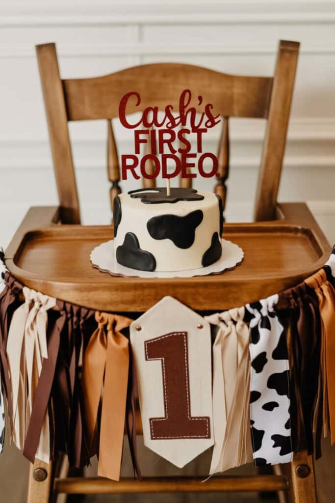 First rodeo birthday smash cake with cow print design and red “Cash’s First Rodeo” cake topper on a decorated high chair with western bandana banner for a cowboy first birthday party.