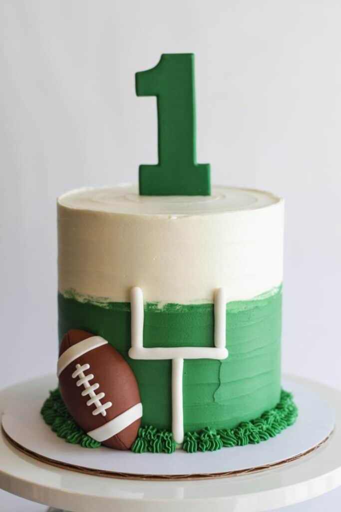 Football-themed first birthday smash cake with green frosting shaped like a field, white goal post details, a fondant football, and a green number one topper.
