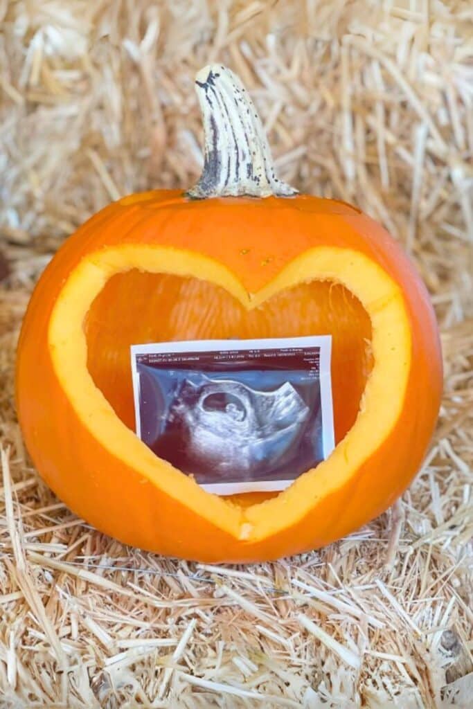 Ultrasound photo displayed inside a pumpkin with a heart-shaped cutout, placed on hay for a fall-themed pregnancy announcement.