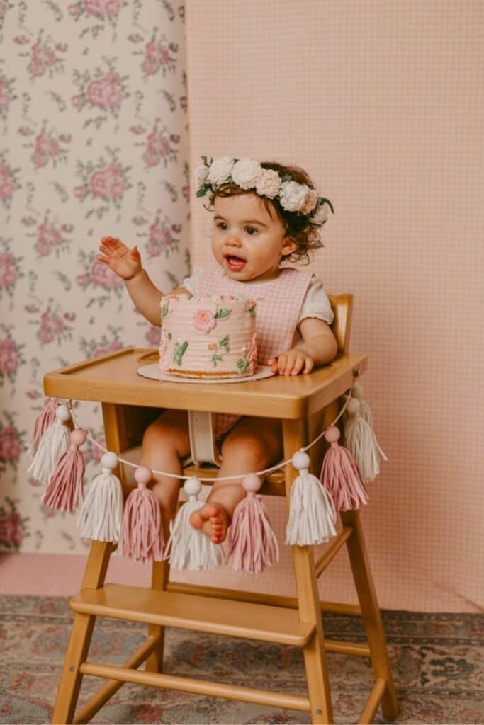 One Silly Goose birthday party decorations for girl featuring a pink floral smash cake, baby in high chair with pink and white tassel garland, soft pink gingham backdrop, flower crown headband, and vintage-inspired first birthday photo setup