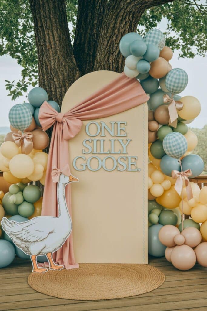 One Silly Goose birthday photo backdrop with pastel balloon arch, custom sign, blush fabric draping, and goose cutout for girl first birthday party