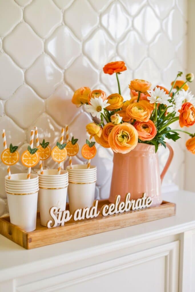 Citrus-themed baby shower drink station with stacked white cups, orange slice drink toppers, a peach vase of orange ranunculus and daisies, and a “Sip and celebrate” sign—perfect for a little cutie baby shower.