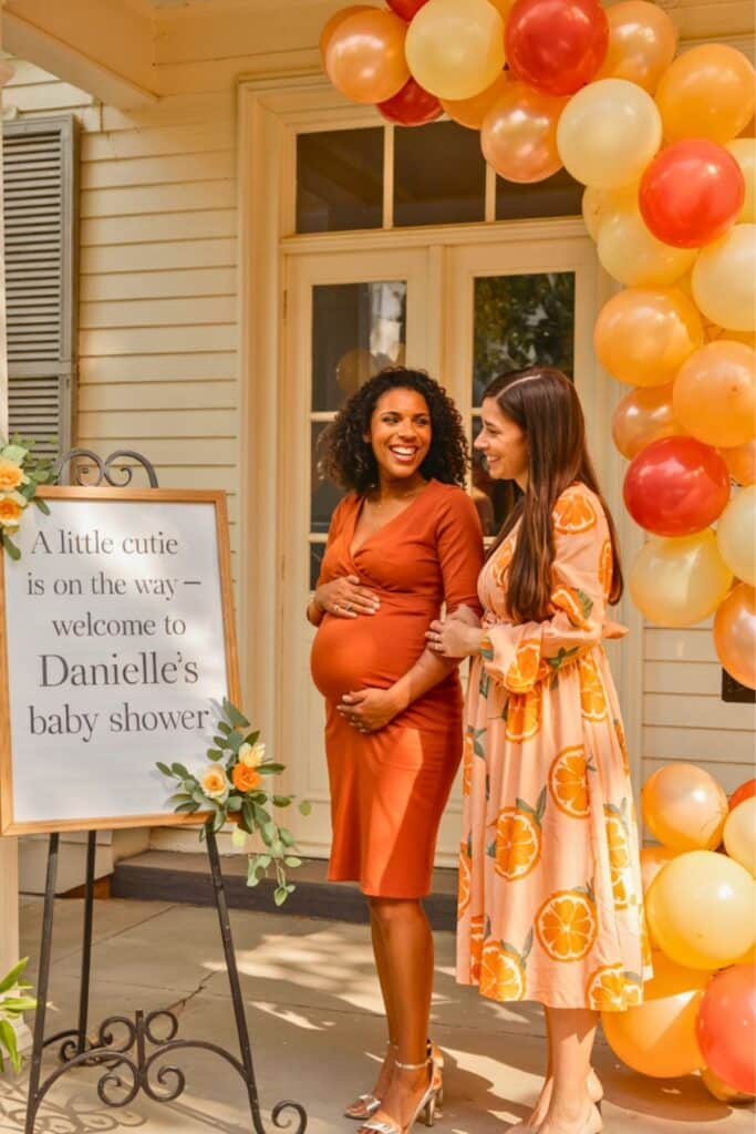 Pregnant woman standing next to a personalized “A little cutie is on the way” baby shower welcome sign with a friend, surrounded by orange, yellow, and red balloons—perfect entrance setup for a cutie orange baby shower theme.