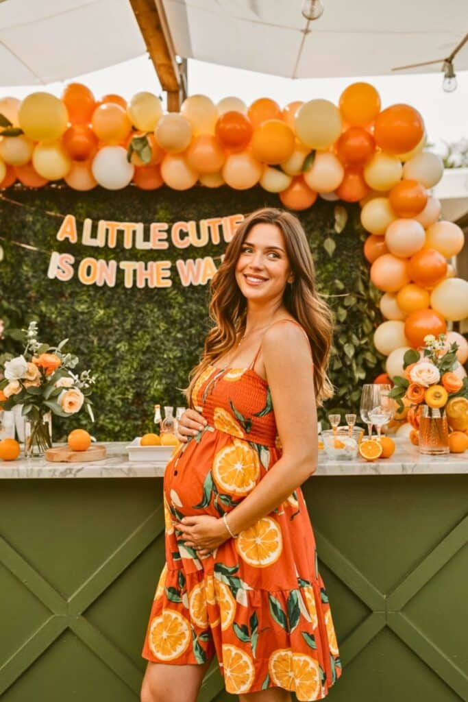 Pregnant woman at a cutie orange baby shower, standing in front of a citrus-themed balloon garland and greenery backdrop with the sign “A Little Cutie Is On The Way.” The setup features orange baby shower decorations, fresh oranges, floral centerpieces, and a green bar, perfect for a summer baby shower theme.