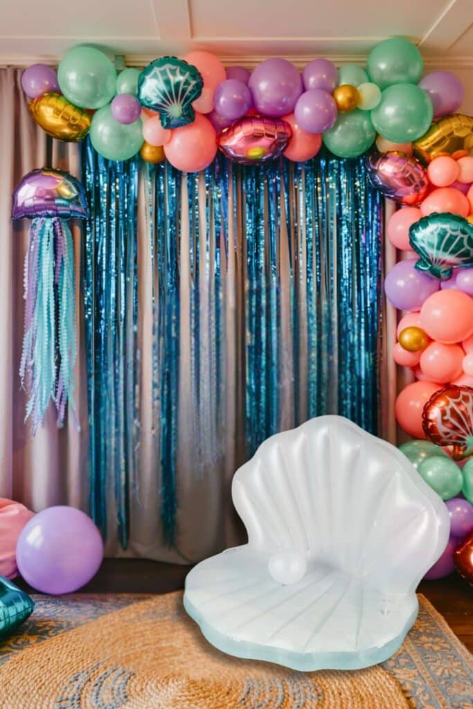 Mermaid birthday party backdrop with pastel balloon garland, metallic jellyfish balloons, blue fringe curtains, and an inflatable seashell chair; perfect for an under-the-sea photo booth setup.