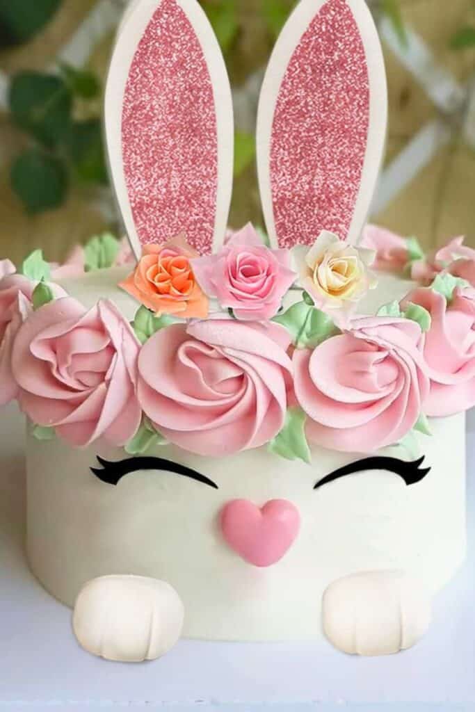 Bunny birthday cake with pink glitter ears, buttercream rose flower crown, heart nose, closed eyes, and fondant paws—“Somebunny Is Three” cake idea.