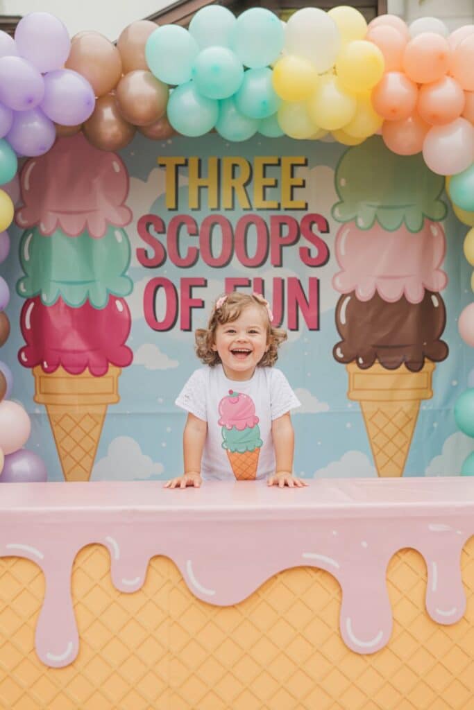Ice cream–themed 3rd birthday party backdrop reading “Three Scoops of Fun,” with rainbow balloon garland, pastel waffle table with pink drip, and a smiling toddler in an ice-cream shirt—sweet summer third birthday girl party décor and photo spot.