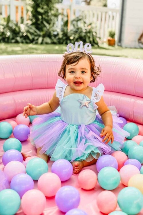 Baby girl celebrating her first birthday in a pastel “Oneder the Sea” theme, wearing a mermaid-inspired tutu dress and “one” crown, sitting in a pink inflatable ball pit filled with teal, lavender, and pink balls—perfect for an under the sea first birthday party.