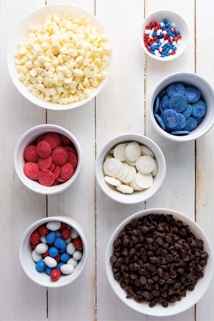 Overhead view of ingredients for patriotic chocolate bark, including bowls of dark chocolate chips, white chocolate chips, red and blue candy melts, star sprinkles, and red, white, and blue M&M-style candies on a white wood background. Perfect for a festive 4th of July dessert.  