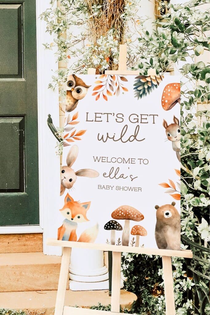 Woodland baby shower welcome sign on an easel with watercolor forest animals, mushrooms, and the phrase “Let’s Get Wild – Welcome to Ella’s Baby Shower,” displayed outside near a green front door.