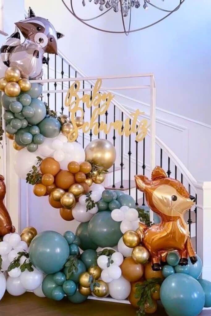 Woodland baby shower balloon arch with forest green, white, gold, and rust-colored balloons, decorated with raccoon and deer foil balloons and a gold script sign that says “Baby Schwartz,” displayed indoors near a staircase.