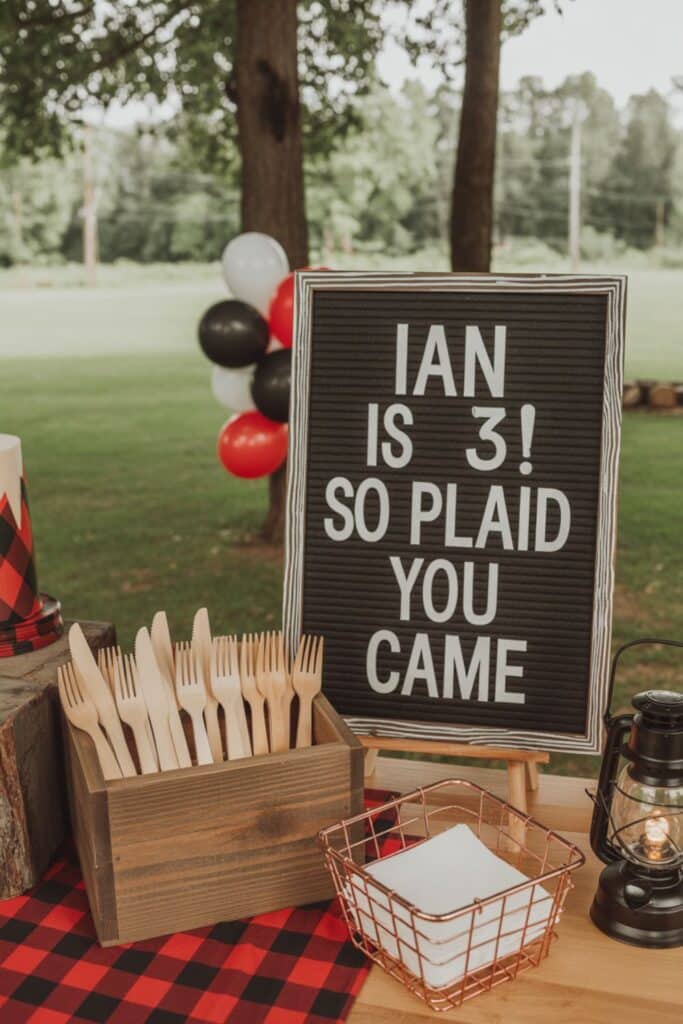 Rustic lumberjack-themed 3rd birthday party table with plaid tablecloth, wooden utensils, lantern, and a letter board sign that reads “Ian is 3! So plaid you came.”