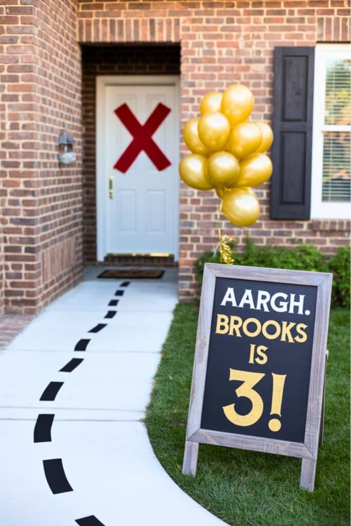 Pirate-themed 3rd birthday party entrance with a chalkboard sign that says "Aargh. Brooks is 3!" next to gold balloons and a red X on the door. A dotted path leads to the party for a fun treasure hunt vibe.