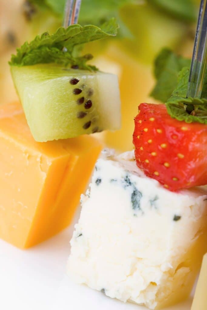 Close-up of fruit and cheese appetizer skewers with kiwi, strawberry, cheddar, blue cheese, and mint. A colorful and refreshing party snack perfect for baby showers, summer parties, or a light appetizer spread.