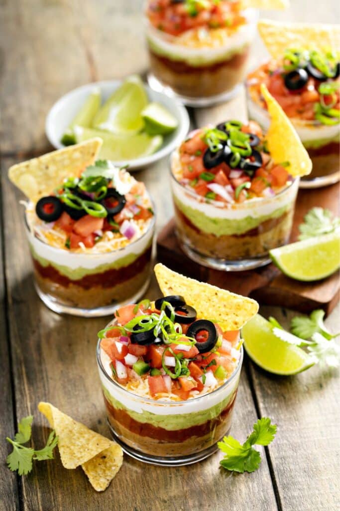Individual 7-layer dip cups filled with beans, guacamole, sour cream, salsa, cheese, olives, and tomatoes, topped with green onions and a tortilla chip. A colorful and fun appetizer idea perfect for birthday parties, baby showers, or summer gatherings.
