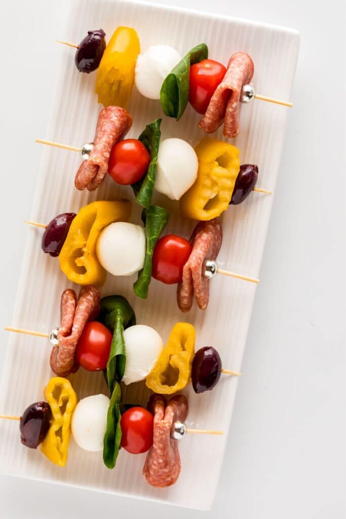 Antipasto skewers with salami, mozzarella, cherry tomatoes, spinach, olives, and bell peppers, arranged on a white platter. A colorful and crowd-pleasing party appetizer perfect for baby showers, birthday parties, or holiday entertaining.