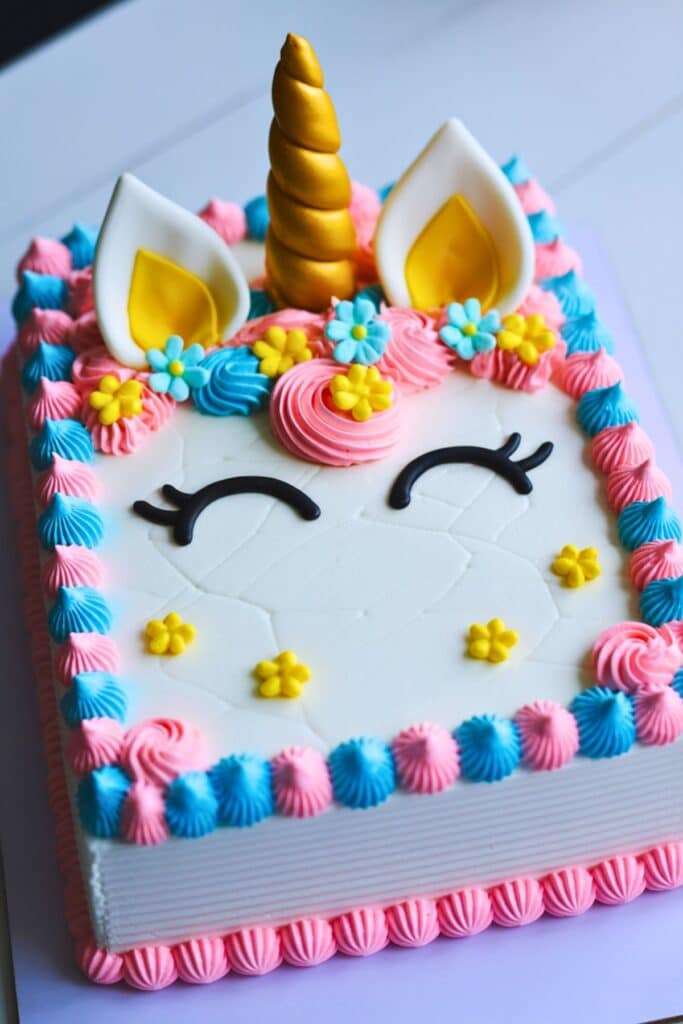 Rectangular unicorn birthday cake decorated with pink, blue, and yellow frosting, a gold fondant horn, fondant ears, and floral accents — a colorful and playful dessert for a unicorn themed celebration.