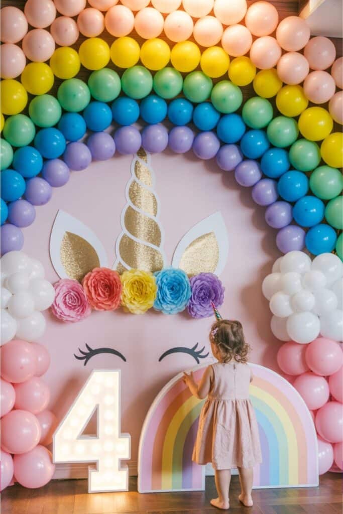 Toddler in a pink dress standing in front of a rainbow balloon arch and unicorn backdrop with gold glitter details, oversized flowers, a pastel rainbow prop, and a light-up number 4 — a vibrant setup for a unicorn-themed fourth birthday party.