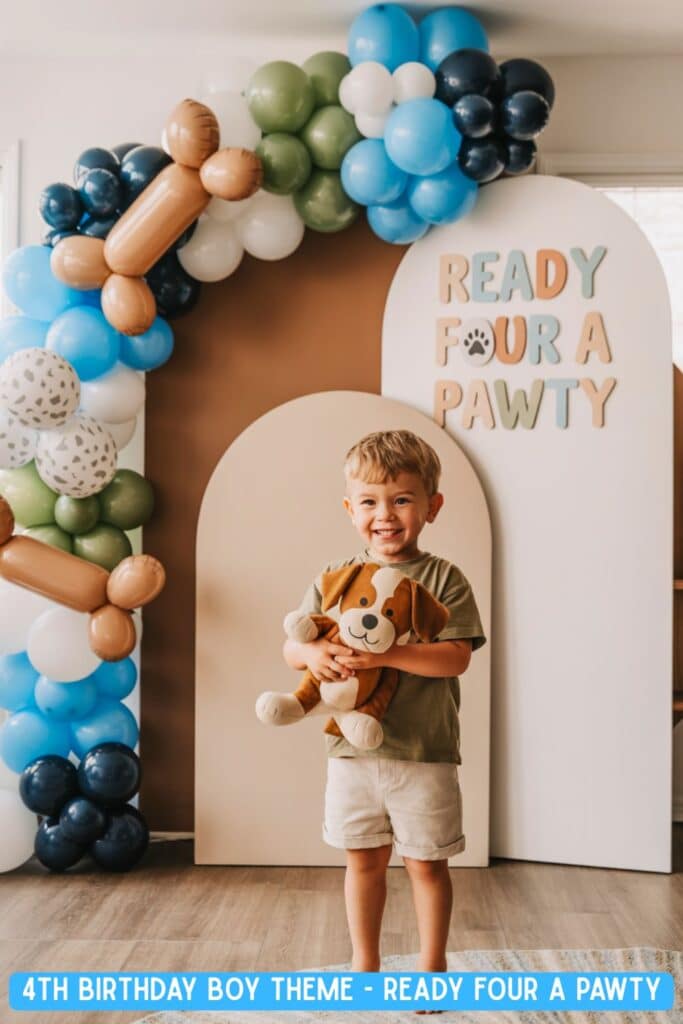 Little boy smiling at his puppy-themed 4th birthday party, holding a stuffed dog in front of a “Ready FOUR a Pawty” sign with balloon arch featuring dog bone balloons in brown, blue, green, and white. Cute dog birthday party for a four-year-old boy.
