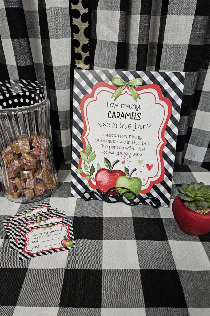 Apple of My Eye baby shower caramel guessing game with red and green apple sign, gingham background, and jar of caramels — fall baby shower game station idea.