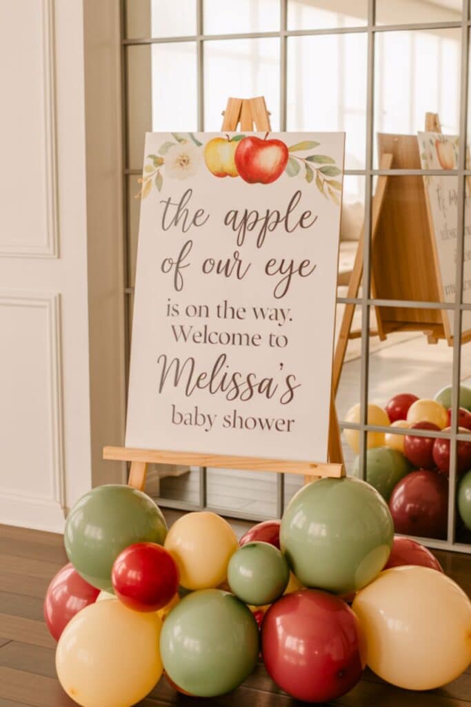 Apple of Our Eye baby shower welcome sign on an easel with red, green, and cream balloon garland, perfect for a fall-themed baby shower.