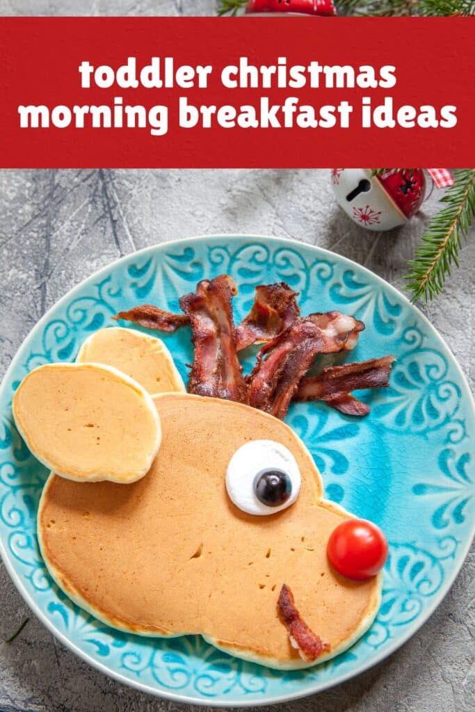 A festive Christmas breakfast for toddlers featuring a reindeer-shaped pancake with bacon antlers, a whipped cream and blueberry eye, and a cherry tomato nose. This kid-friendly Christmas morning breakfast idea is served on a decorative blue plate with pine sprigs and jingle bells in the background.