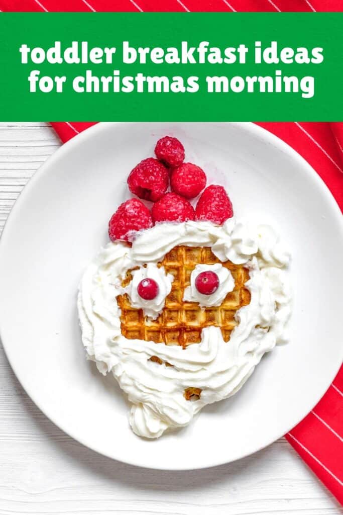 A Santa-themed waffle toddler breakfast served on a white plate, decorated with whipped cream for the beard and hat trim, raspberries for Santa's hat, and two red cranberries as eyes. This fun and festive Christmas morning breakfast idea for toddlers is playful, sweet, and perfect for getting little ones excited to eat.