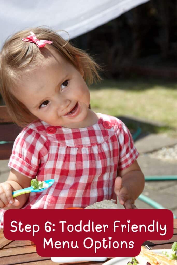 Toddler girl in a red checkered dress eating toddler-friendly food at her outdoor second birthday party.