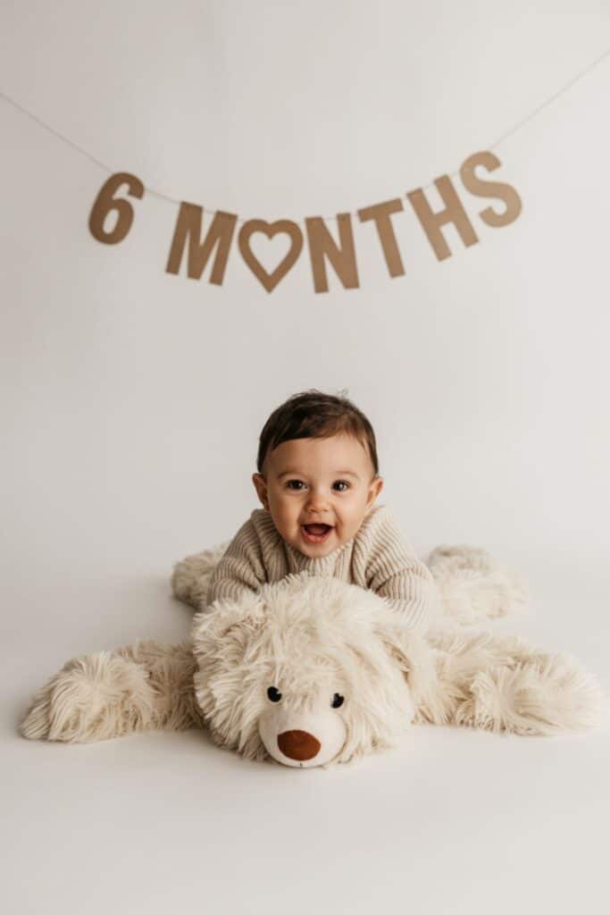 Six month milestone photo with baby on giant teddy under “6 Months” banner.
