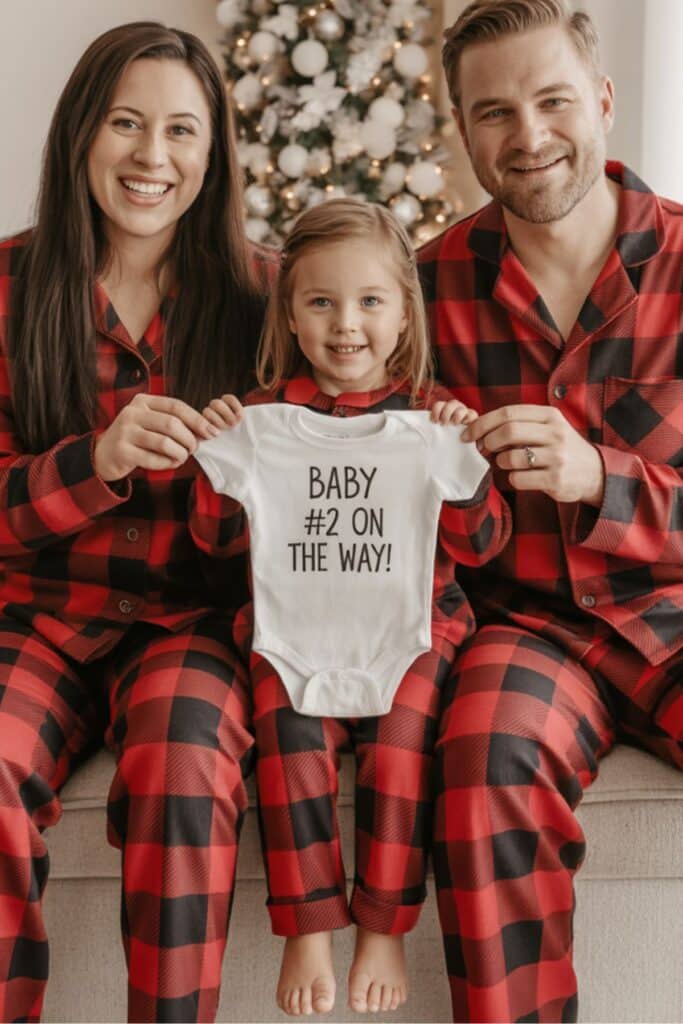 Christmas pregnancy announcement photo with parents and daughter in matching red plaid pajamas holding a baby onesie that reads “Baby #2 on the way!” in front of a decorated Christmas tree, perfect holiday reveal idea for announcing a second pregnancy.