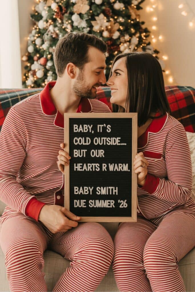 Couple in matching red striped Christmas pajamas sitting in front of a decorated Christmas tree, holding a letterboard that reads “Baby, it’s cold outside… but our hearts are warm. Baby Smith due Summer ’26” for a cozy holiday pregnancy announcement photo.
