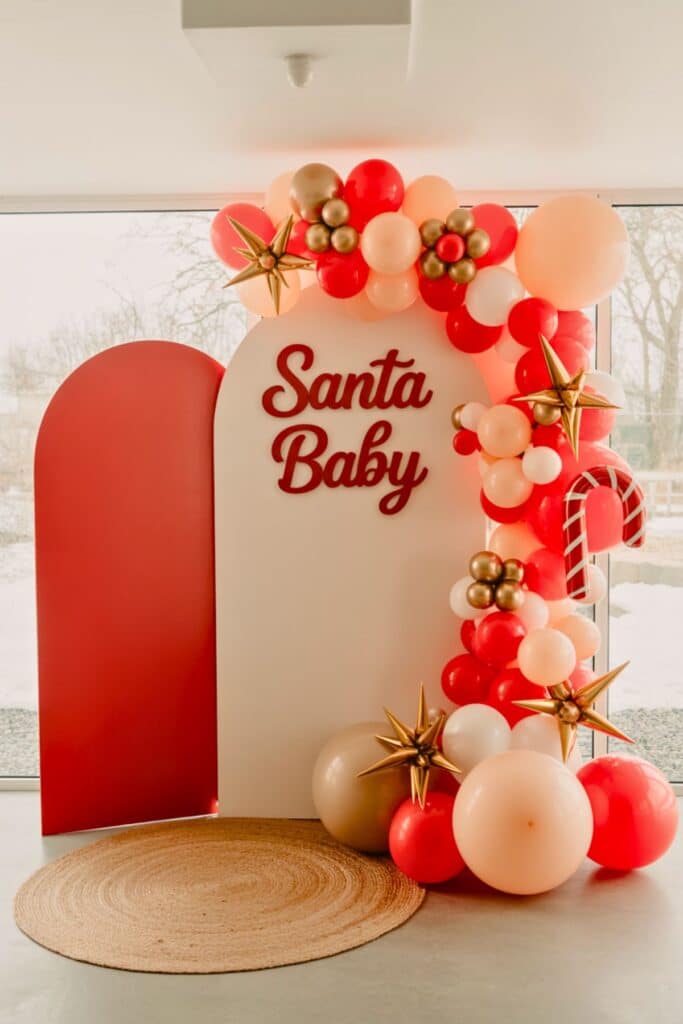 Red and white and pink Santa Baby baby shower balloon arch with gold star accents, candy cane decor, and festive Christmas backdrop — perfect inspiration for a December or Christmas-themed baby shower.