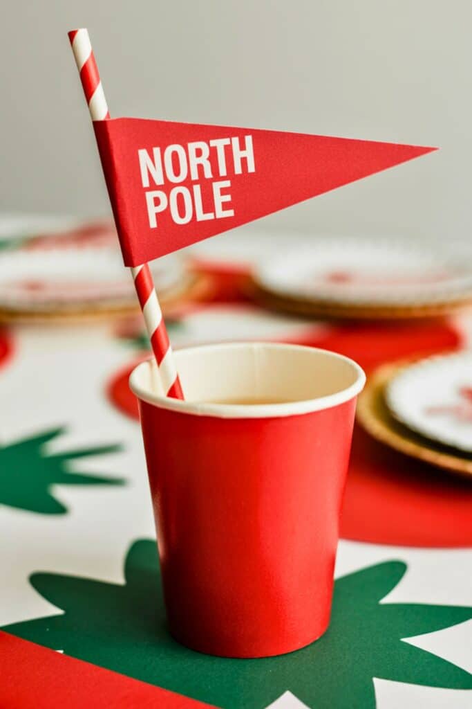 Red paper cup with red-and-white striped straw topped with a small red flag reading “North Pole,” set on a Christmas-themed table with green and red cutouts and festive holiday plates — perfect detail idea for a Santa Baby or Christmas baby shower.