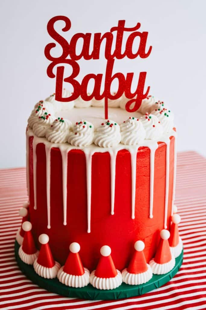 Red and white Santa Baby baby shower drip cake with white frosting swirls topped with sprinkles, glittery red “Santa Baby” topper, and mini Santa hat decorations around the base — a festive dessert centerpiece for a Christmas-themed baby shower.