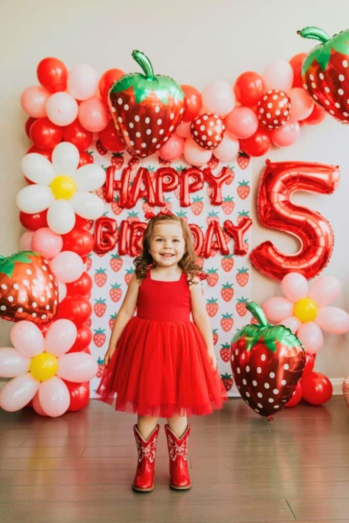 A smiling little girl wearing a red tutu dress and red cowboy boots stands in front of a vibrant strawberry-themed 5th birthday party backdrop. The colorful decorations include red and pink balloons, large strawberry foil balloons, daisy balloon flowers, and a big number 5 balloon, creating a cheerful and whimsical party scene perfect for a “Berry Sweet” fifth birthday celebration.
