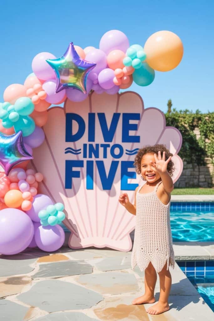 Little girl celebrating her 5th birthday with a pastel balloon arch and a giant shell backdrop that reads “Dive Into Five,” standing by the pool on a sunny day.