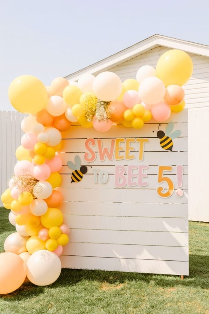 A “Sweet to Bee 5” birthday party backdrop featuring a white shiplap wall with playful pastel letters, cute bee graphics, and a vibrant balloon garland in shades of yellow, peach, blush pink, and white. The whimsical outdoor setup is perfect for a bee-themed 5th birthday party for girls.