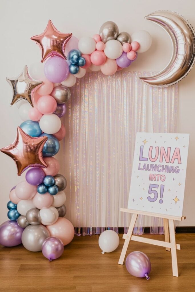 Balloon arch in pink, purple, silver, and blue tones with metallic star and moon balloons beside a sign that reads “Luna Launching Into 5!” against a shimmering iridescent curtain backdrop.