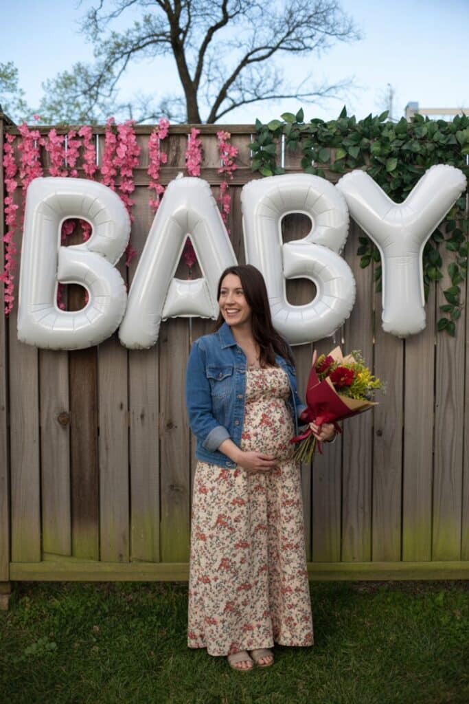 Pregnant woman at a spring baby shower standing under large silver “BABY” letter balloons on a wooden fence with pink florals and greenery, holding a bouquet in a floral dress and denim jacket.