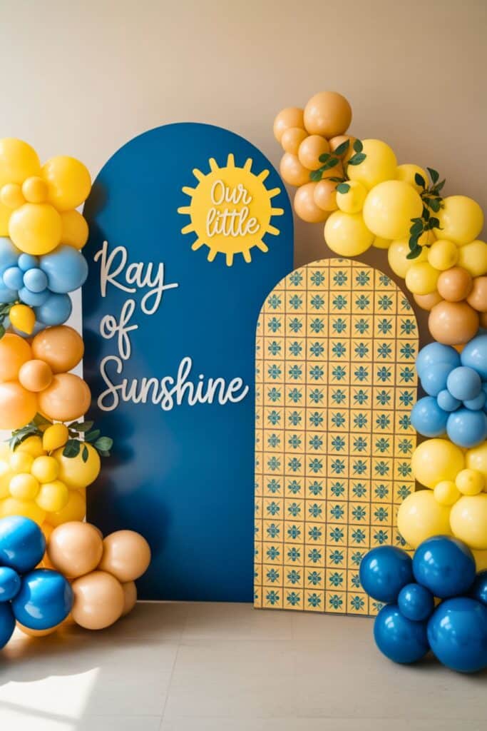 ‘Our little ray of sunshine’ baby shower backdrop in navy and yellow with sunshine cutout, patterned panel, and yellow–blue balloon garland.