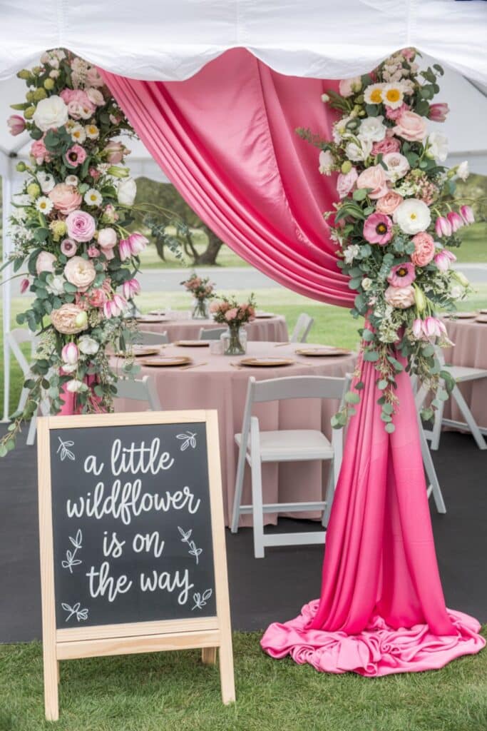 A little wildflower is on the way’ chalkboard under a pink draped entry with lush floral pillars at an outdoor spring baby shower.