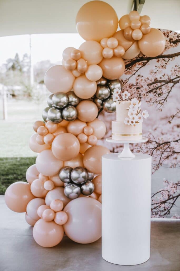 Cherry blossom Spring Baby Shower backdrop with a blush balloon arch and white pedestal holding a two-tier cake topped with sugar blossoms.