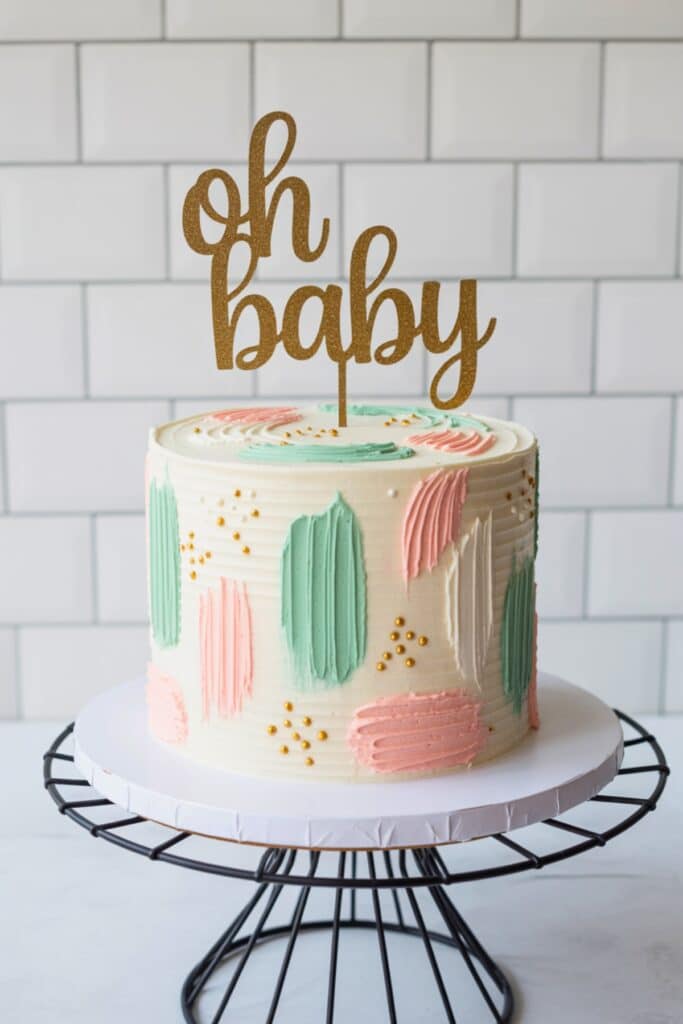 Gender reveal cake with “oh baby” topper, neutral buttercream, palette-knife strokes in blush and sage, and gold sprinkle accents.