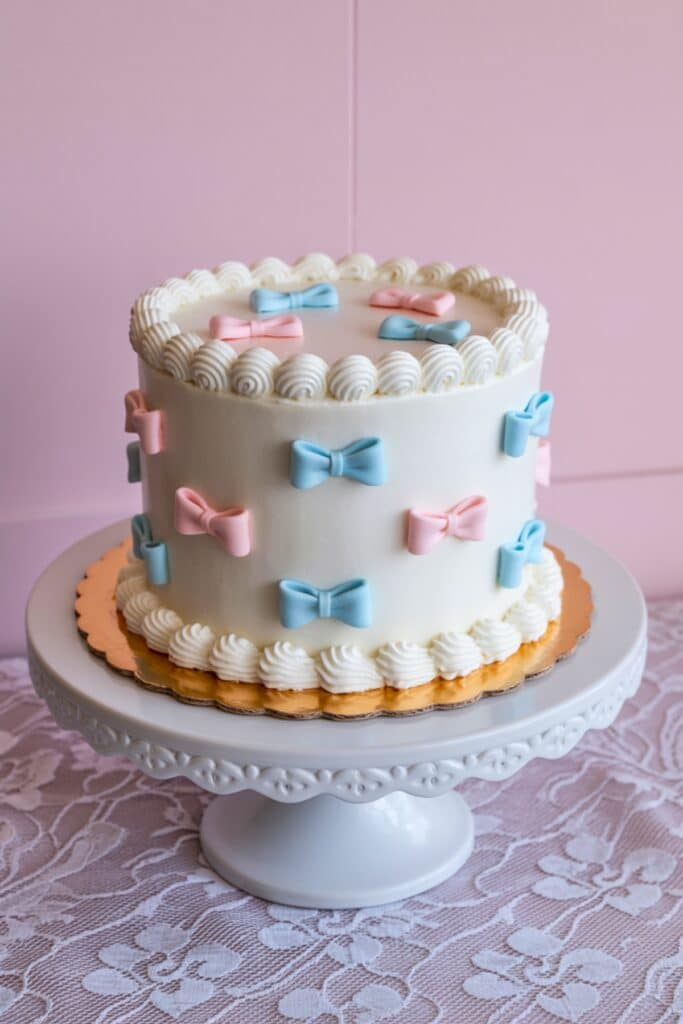 Pink and blue bows gender reveal cake on lace table—clean white frosting with piped shell border; stylish, easy design for bakers.