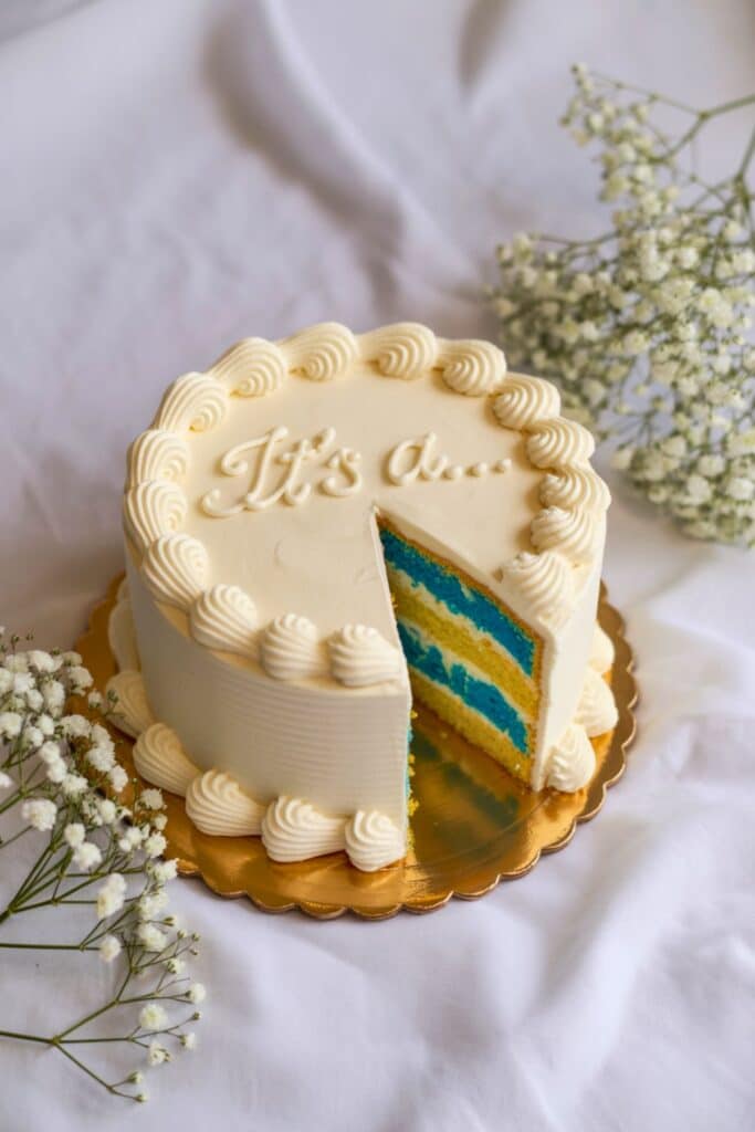 Gender reveal cake with “It’s a…” piped on top, smooth white buttercream, and a slice showing bright blue cake layers; baby’s breath decor.