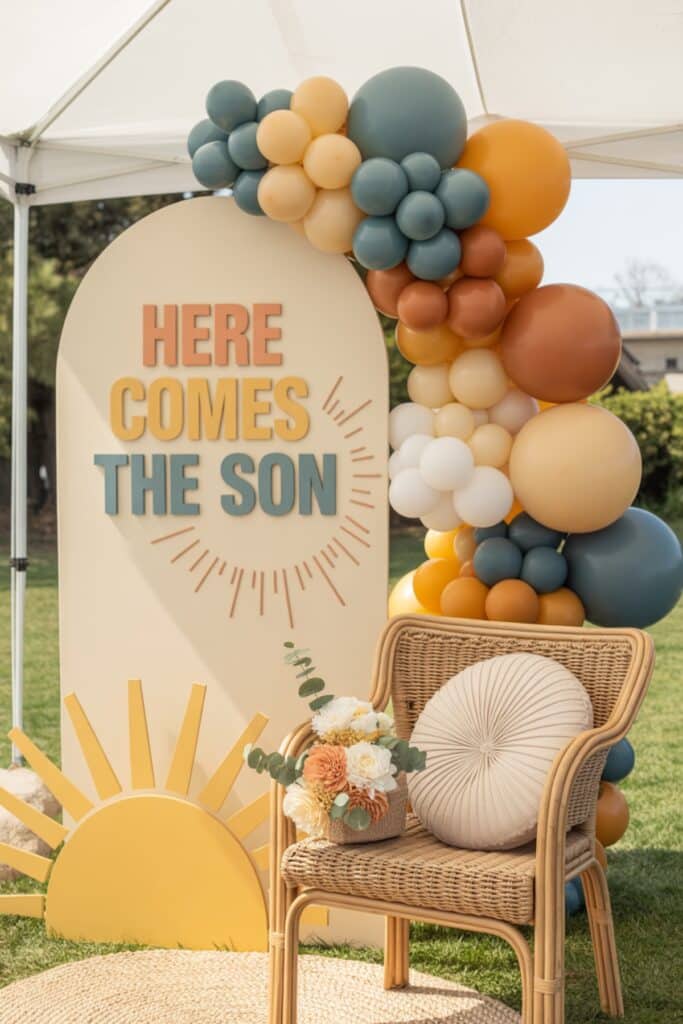 Outdoor Here Comes the Son baby shower seating area with a retro sunrise backdrop sign, warm-toned balloon garland, rattan chair, and soft neutral floral arrangement.