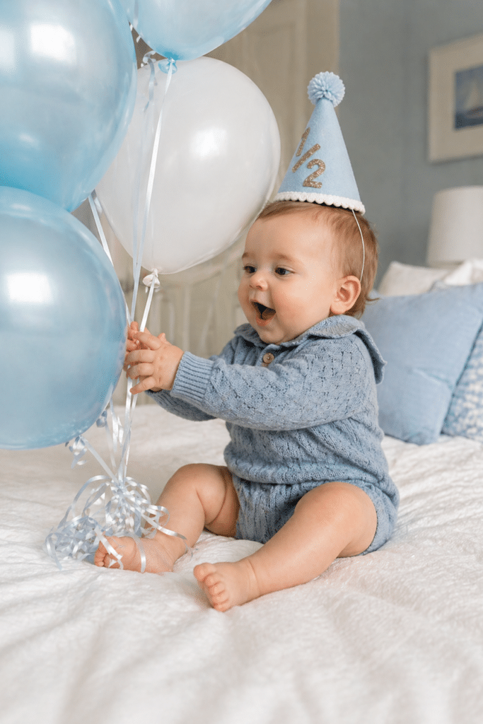 baby boy sitting on bed with blue and white balloons wearing 1/2 hat, easy at home half birthday photo idea
