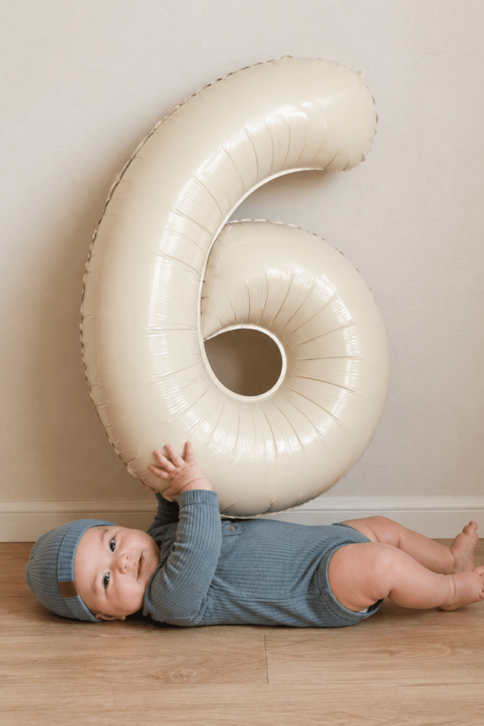 baby boy lying on floor holding large number 6 balloon, simple half birthday photo idea for 6 month milestone