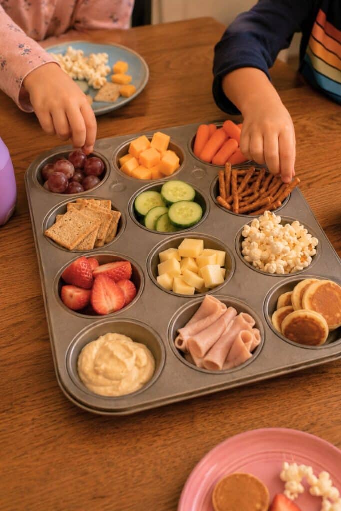 Muffin tin snack board dinner for kids with popcorn, cheese cubes, crackers, fruit, mini pancakes, pretzels, veggies, and turkey roll-ups arranged in individual sections on a wooden table. Two children reach for the snacks during a simple kid-friendly family dinner idea perfect for picky eaters and toddlers.