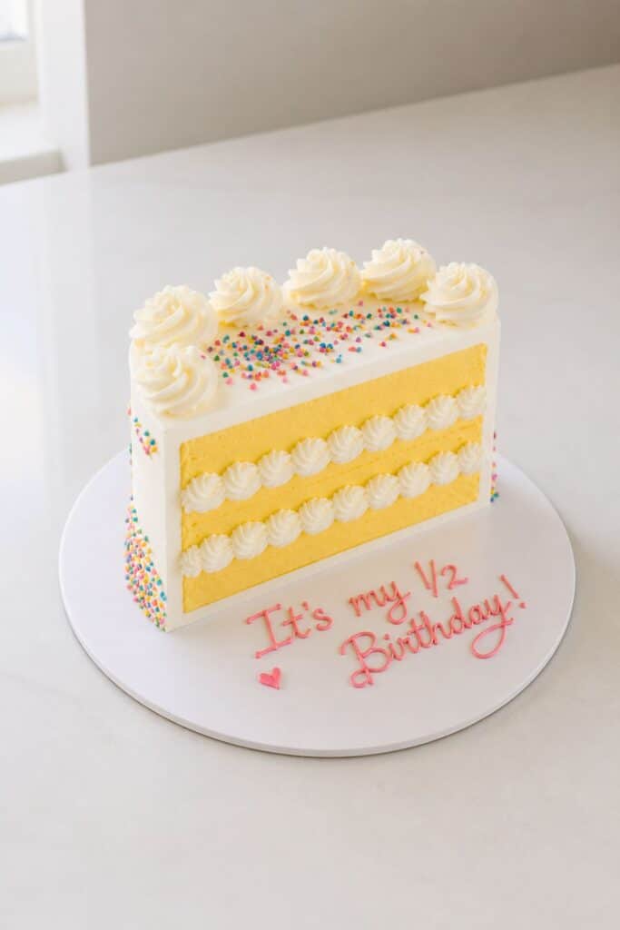 Half birthday cake designed like a slice of vanilla layer cake with rainbow sprinkles and “It’s my 1/2 Birthday!” written in pink frosting for a cute six month birthday party idea.